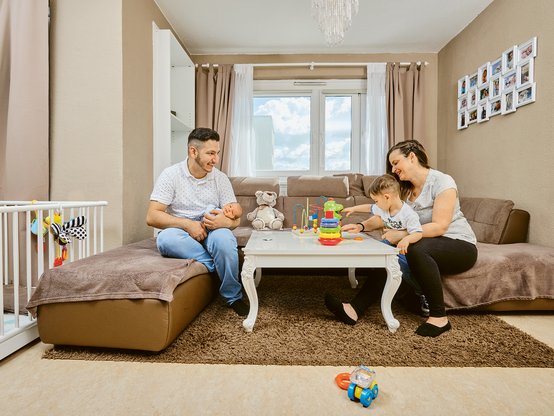 Innenaufnahme: In einem Wohnzimmer sitzen vier Personen - zwei Erwachsene, ein Kleinkind und ein Baby - auf einer Couch.