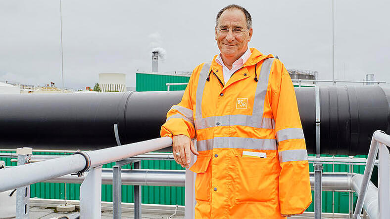 Mann in orangener BSR-Weste steht vor einer Biogasanlage mit großen Röhren im Hintergrund