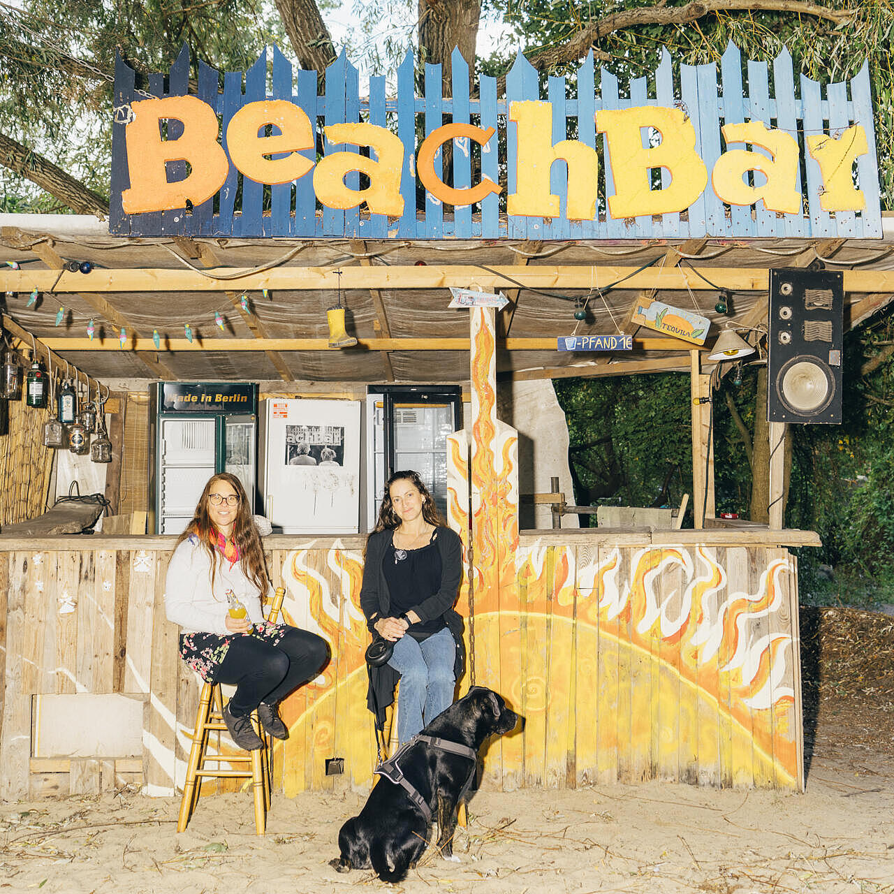 Vor der Beachbar des Wagendorfs sitzen Martina und Irina und Hund. Der Boden ist sandig