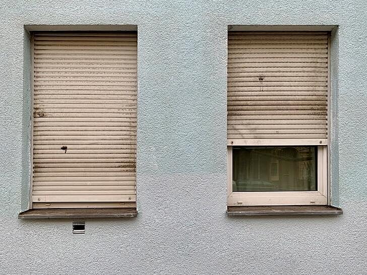 Außenaufnahme: Graue Hausfassade mit zwei Fenstern, bei denen die Rollos komplett (links) bzw. halb herunterlassen sind (rechts)