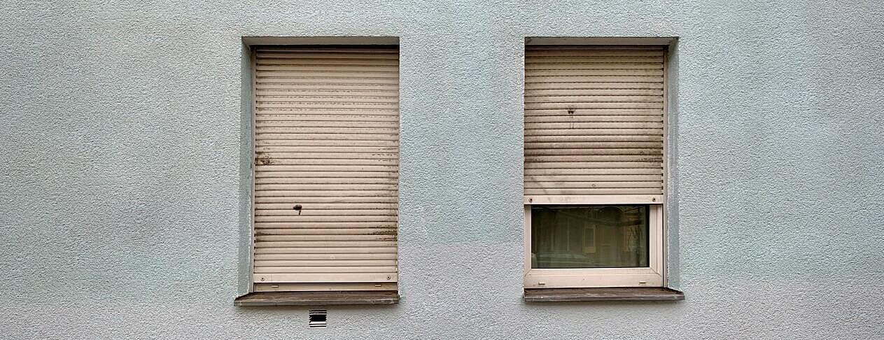 Außenaufnahme: Graue Hausfassade mit zwei Fenstern, bei denen die Rollos komplett (links) bzw. halb herunterlassen sind (rechts)