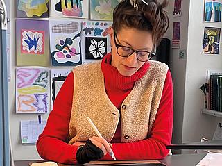 Eine Illustratorin sitzt an ihrem Schreibtisch mit Apple Pen in der Hand. Im Hintergrund sind verschiedene illustrierte Bilder an der Wand.